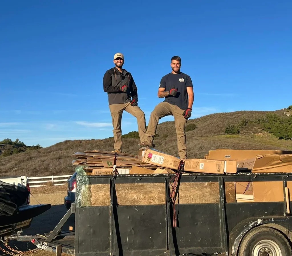 Two BullDawgs team members standing on a loaded junk trailer with cardboard boxes, set against a clear sky and hillside background
