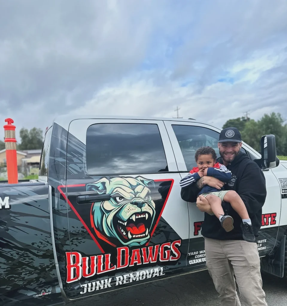 Smiling BullDawgs crew member holding a child in front of their branded junk removal truck on a cloudy day
